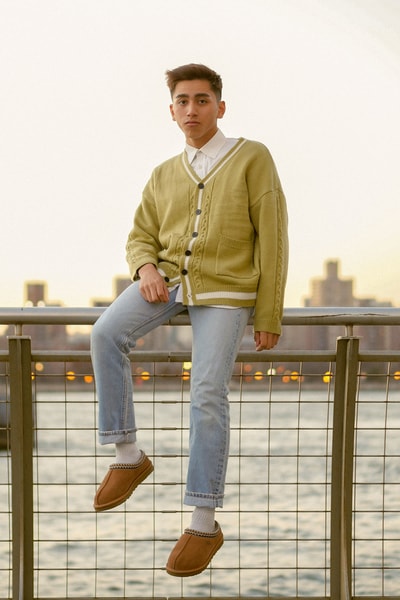 man with green cardigan sitting on rail near river wearing brown ugg shoes